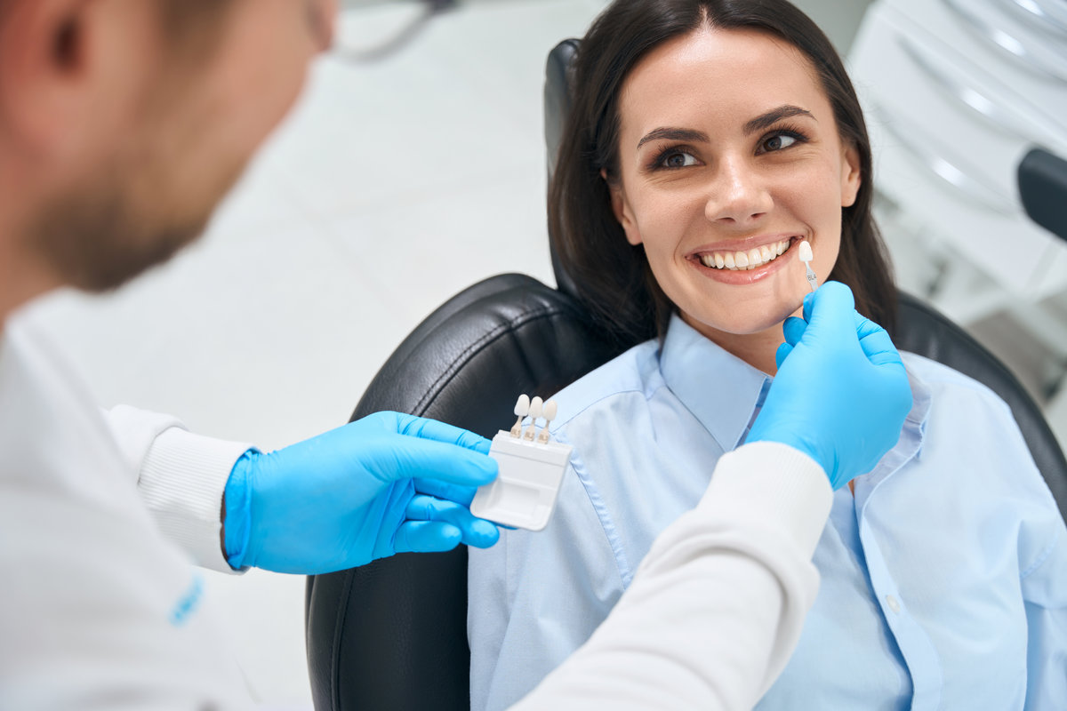 High-qualified dental technician choosing proper size and color of dental crown using some template, smiling woman client sitting in cozy orthodontic chair